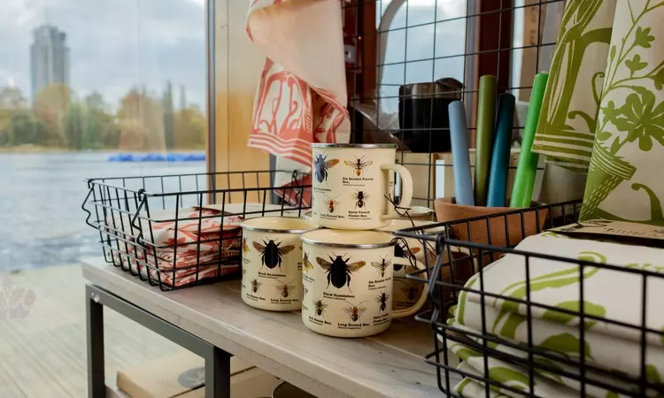 A shop display with the Serpentine lake in the background. A collection on mugs with bee illustrations on sits in the foreground. 